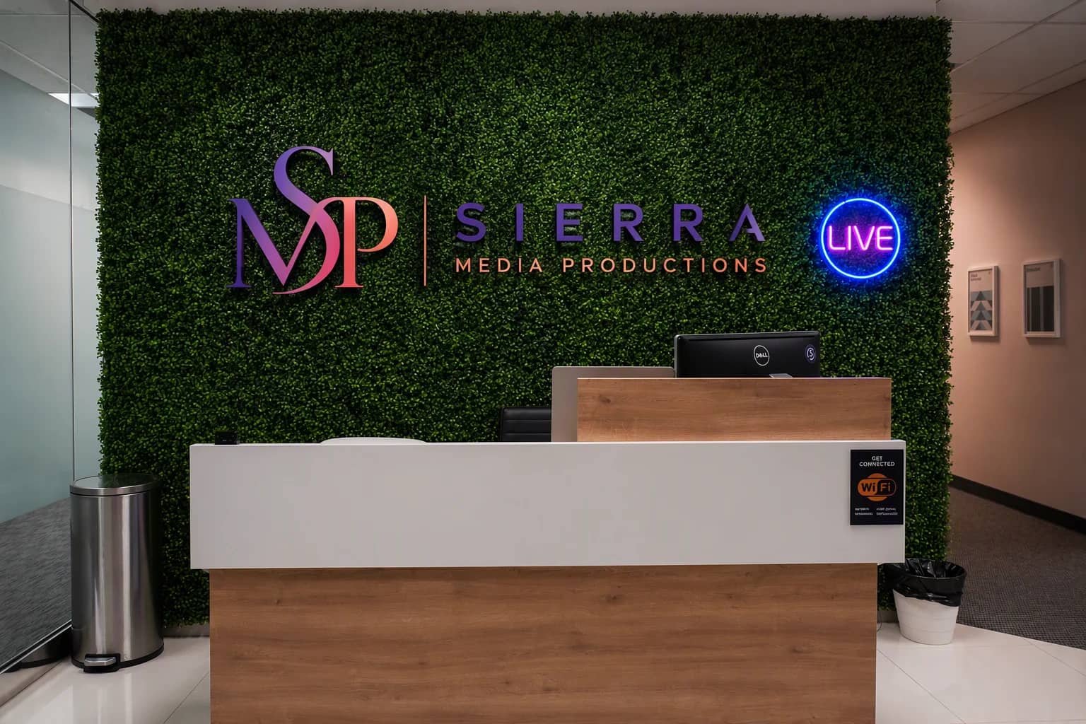 The reception desk and living-greenery wall inside the Sierra Media Productions Dallas podcast studio, with the Sierra Media Productions mark and a LIVE neon sign.