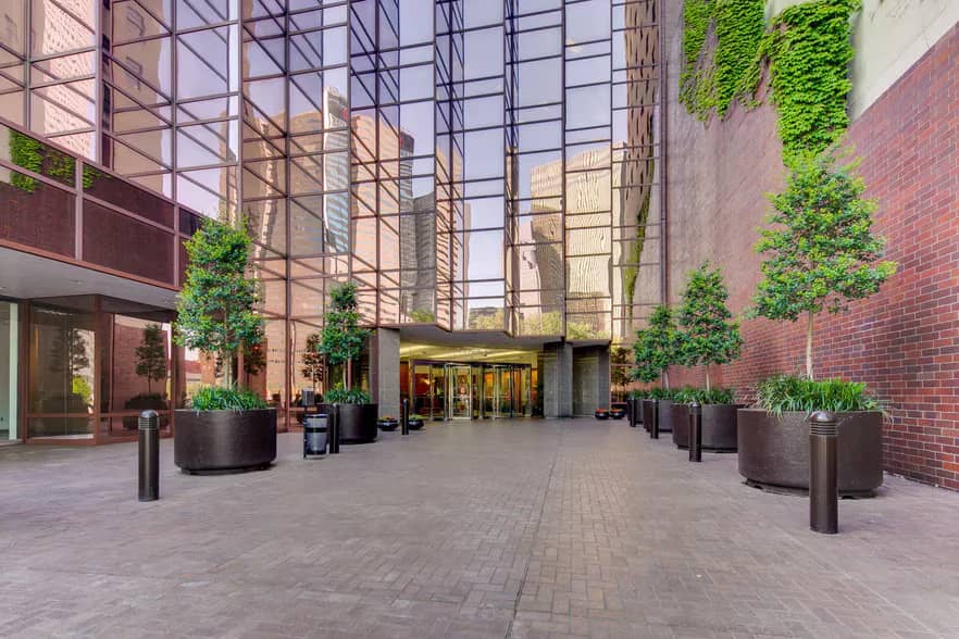 The glass atrium entrance of the downtown Dallas building housing the Sierra Media Productions podcast studio, with an ivy-covered brick wall and planted trees along the paved plaza.